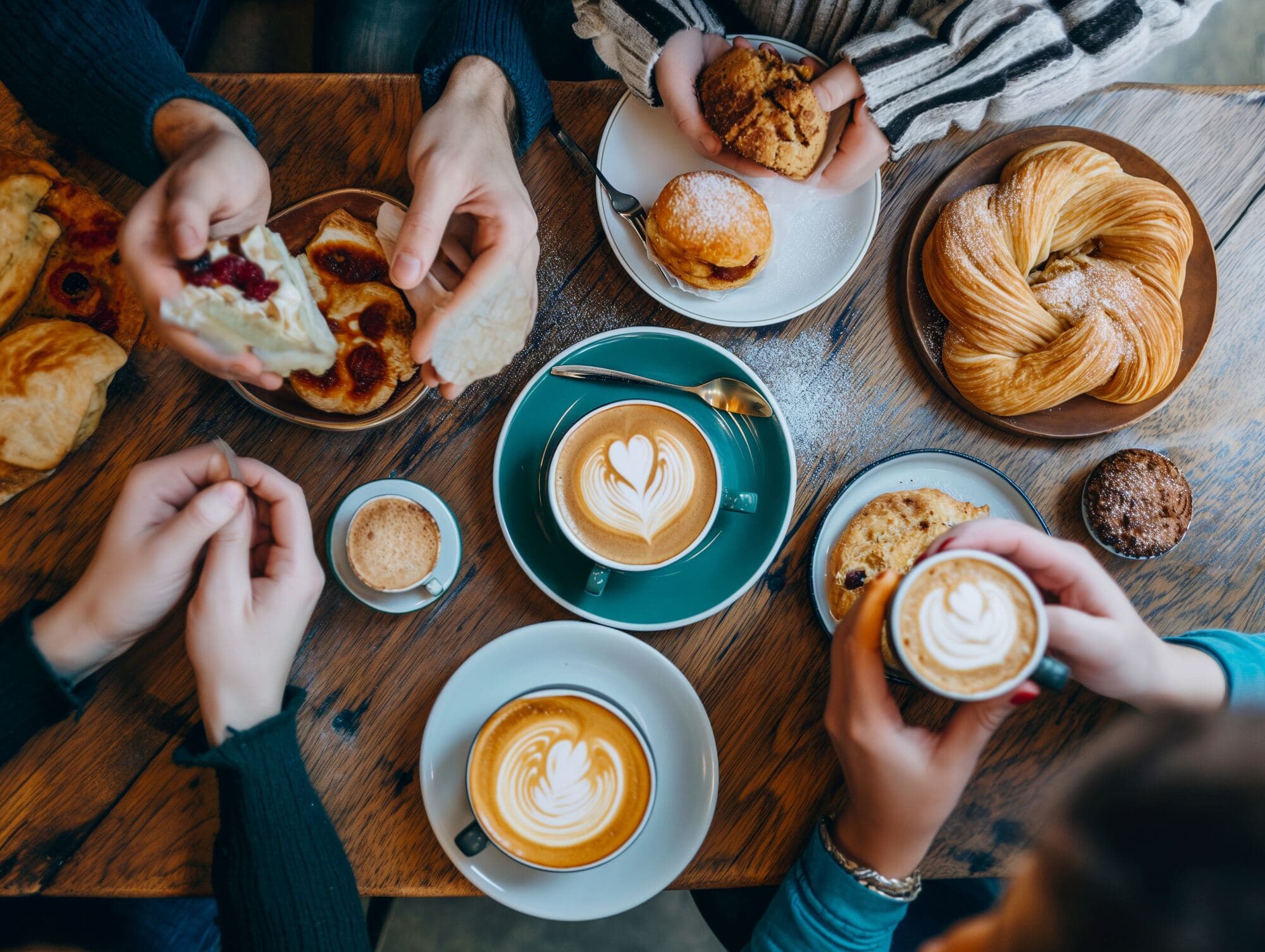 A table of coffees with latte art, pastries and hands reaching in to share them